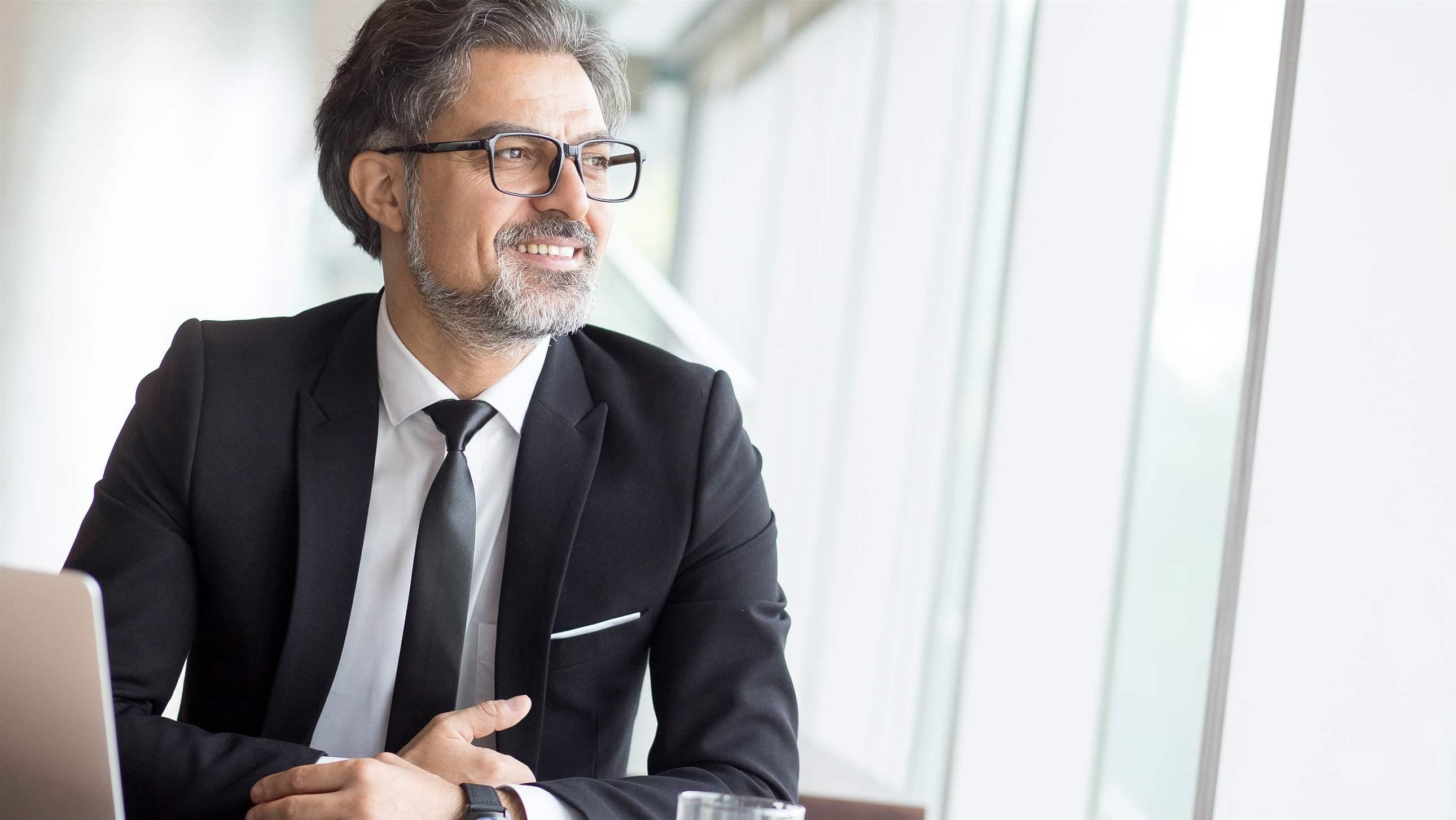 雄心勃勃的高级商人从事新项目。积极成熟的企业家梦想着新的成就。英俊的男人坐在桌子旁，带笔记本电脑看着。商业生活方式概念